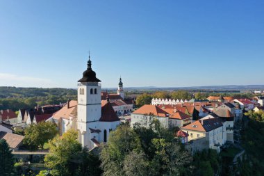 Aerial summer view of Hus Square in Nove Mesto nad Metuji, Czech Republic, featuring a historic church tower, red rooftops, traditional architecture, and clear blue sky.