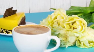 Köpüklü taze espresso kahvesi. Bir fincan kahve, bir buket sarı lale ve açık mavi arka planda fotokopi alanı olan bir dilim pasta. Sevdiğin biri için kahvaltı, 4k