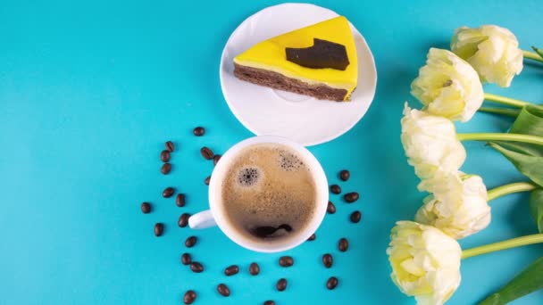 フレッシュアロマエスプレッソコーヒー。黄色のチューリップの花束とコピースペースのある薄い青の背景にケーキのスライスとコーヒーカップ。愛する人のための朝食、 4k — ストック動画