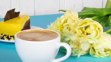Köpüklü taze espresso kahvesi. Bir fincan kahve, bir buket sarı lale ve açık mavi arka planda fotokopi alanı olan bir dilim pasta. Sevdiğin biri için kahvaltı, 4k