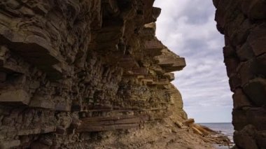 Dalgaların denize vurduğu kayalık bir sahilden kamera hareketi. Arka planda bir pelerin ile