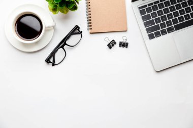 Üst kattaki ofis masasında yatıyordu. Çalışma alanı boş dizüstü bilgisayar, ofis malzemeleri, kalem, yeşil yaprak ve beyaz arka planda kahve fincanı.