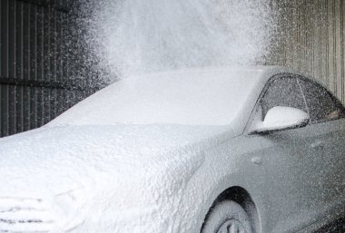 Araba sürücüsü, yüksek basınçlı su püskürten bir hortumla oto yıkamaya daldı. Ön cam ve kaputtaki köpüğü temizledi.