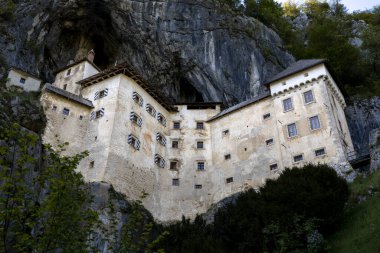 Slovenya 'da yüksek bir kayalık içine inşa edilmiş eşsiz bir ortaçağ kalesi olan tarihi Predjama Şatosu' nun (Predjamski grad) manzarası. Görüntü, bahar aylarında kale mimarisini yürüyen bir patika perspektifinden gösteriyor..