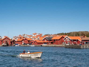 Mollosund, İsveç - 16 Ağustos 2025: Motorlu tekne, İsveç 'in batı kıyısındaki Bohuslan takımadalarında kırmızı ahşap rıhtım evlerini, eski balıkçı kulübelerini ve kayıkhaneleri geçiyor