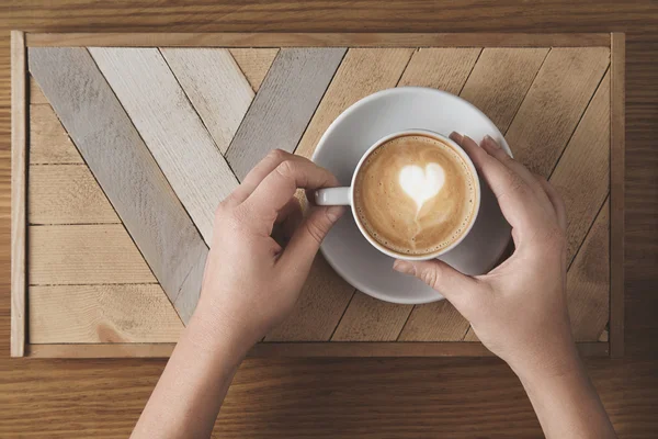 Schöne Frauenhände halten Keramik weiß mit Cappuccino auf einem Holzteller und einem rustikalen Tisch. Milchschaum darüber in Baumform. Draufsicht im Café-Shop. Verkaufspräsentationskonzept. — Stockfoto