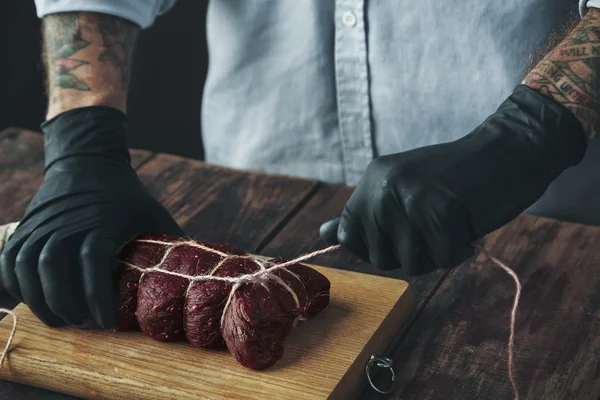 Татуированный мясник вяжет мясо веревкой, чтобы курить деревянный стол — стоковое фото