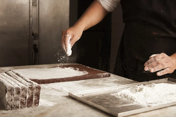 Black man chief pours sugar powder on chocolate cakes before packaging — Stock Photo, Image