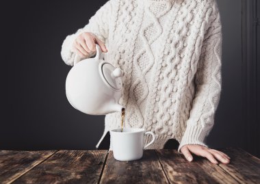 Woman in white sweater pours hot tea from big ceramic teapot