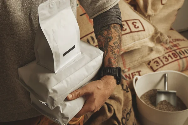 Ο μπάρμαν με τα τατουάζ κρατά κενά πακέτα με καφέ — Φωτογραφία Αρχείου