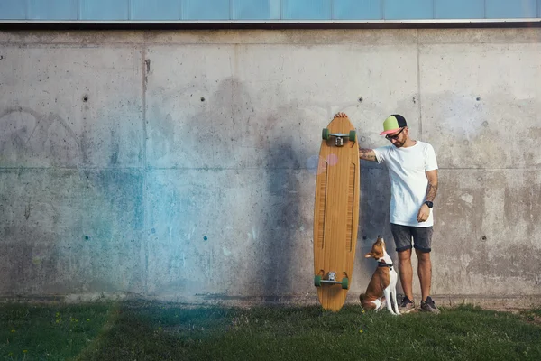 Koira katselee hipsteriä, jolla on longboard — kuvapankkivalokuva