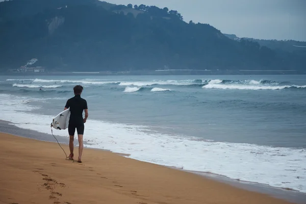 Surffaaja märkäpuvussa kävelee pitkin rantaa — kuvapankkivalokuva