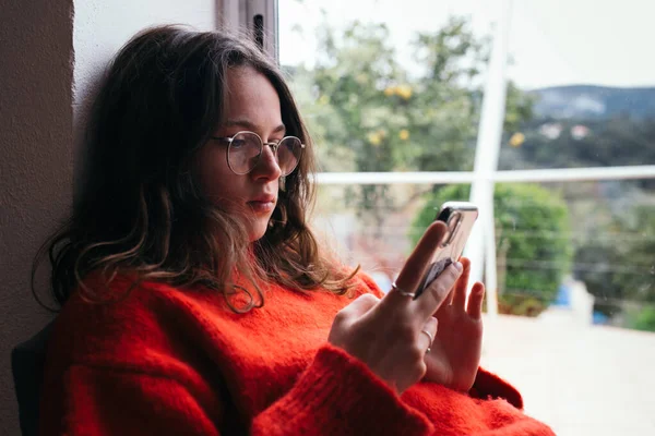 Молода жінка в червоному светрі дивиться на телефон вдома — стокове фото