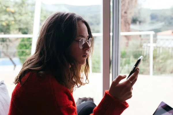 Mujer joven en suéter rojo mira el teléfono en casa —  Fotos de Stock