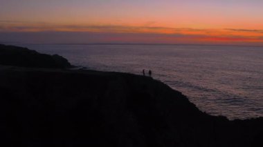 Çiftin silueti gün batımında uçurumun kenarında yürüyor.