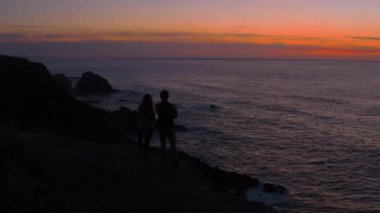 Çiftin silueti gün batımında uçurumun kenarında yürüyor.