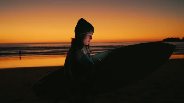 Gün batımının destansı sahilinde sörf tahtası olan genç bir kadın. — Stok video