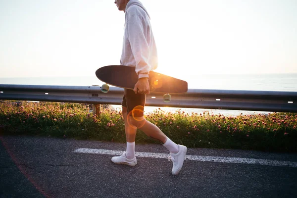 Jovem hipster carrega longboard ao pôr do sol — Fotografia de Stock