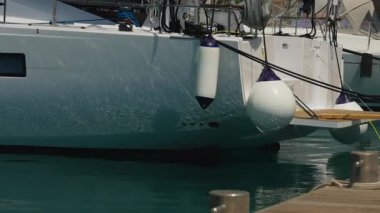 Rıhtımdaki beyaz gövdeyi koruyan tekne çamurlukları, çamurluk halatları ve Cleat düğümleri görünür, deniz kenarındaki güneş ışığı ve nazik yansımalarla bakım havası.