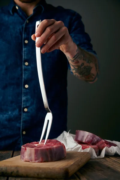 Tatuado carnicero mano sostiene tenedor de carne en filete fresco para la cena —  Fotos de Stock