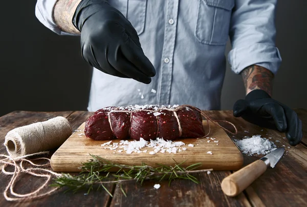 Tattoed butcher in black gloves salts tied piece of meat to smoke — Stock Photo, Image