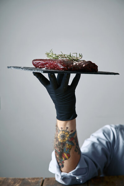 Tattoed hand in black glove holds stone plate with steak