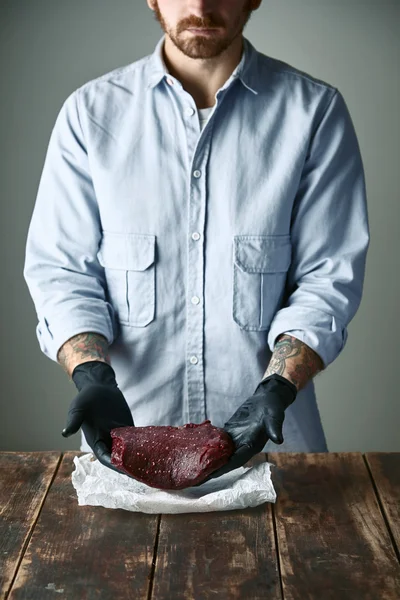 Бородатый мясник-хипстер в черных перчатках предлагает кусок мяса кита — стоковое фото