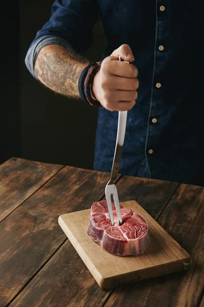 Carnicero mano sostiene tenedor de carne en carne cruda —  Fotos de Stock