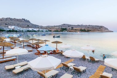 Boş bir sahil beldesinde şemsiyeler ve güneşlikler - Ege ve Akdeniz 'deki Yunanistan adalarında tatil kavramı. Rodos 'un doğu kıyısındaki Charaki adlı küçük bir köyde kapalı bir körfezde sahil.