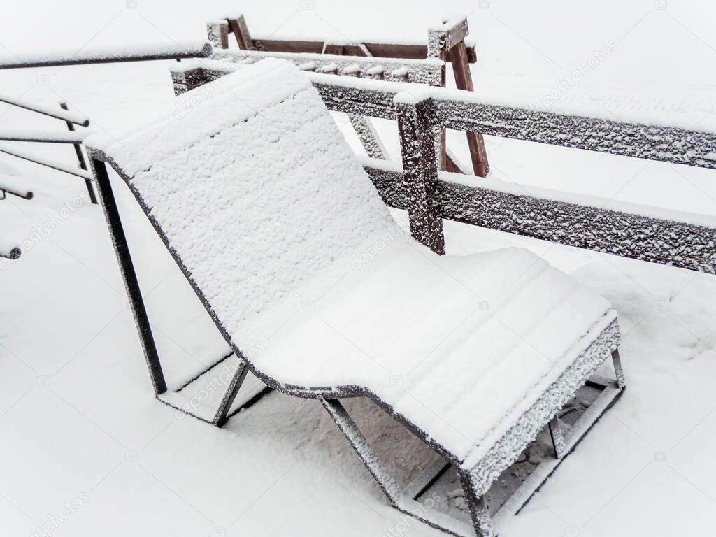 A sun lounger covered with snow stands outside on a cloudy winter day