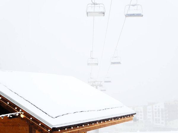 The roof of a wooden house covered with snow on the background of a chairlift in the fog