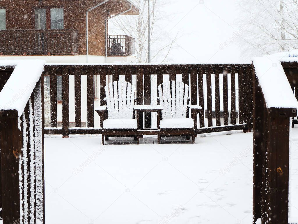 Two wooden chairs connected to each other stand on the terrace covered with snow in winter during a snowfall