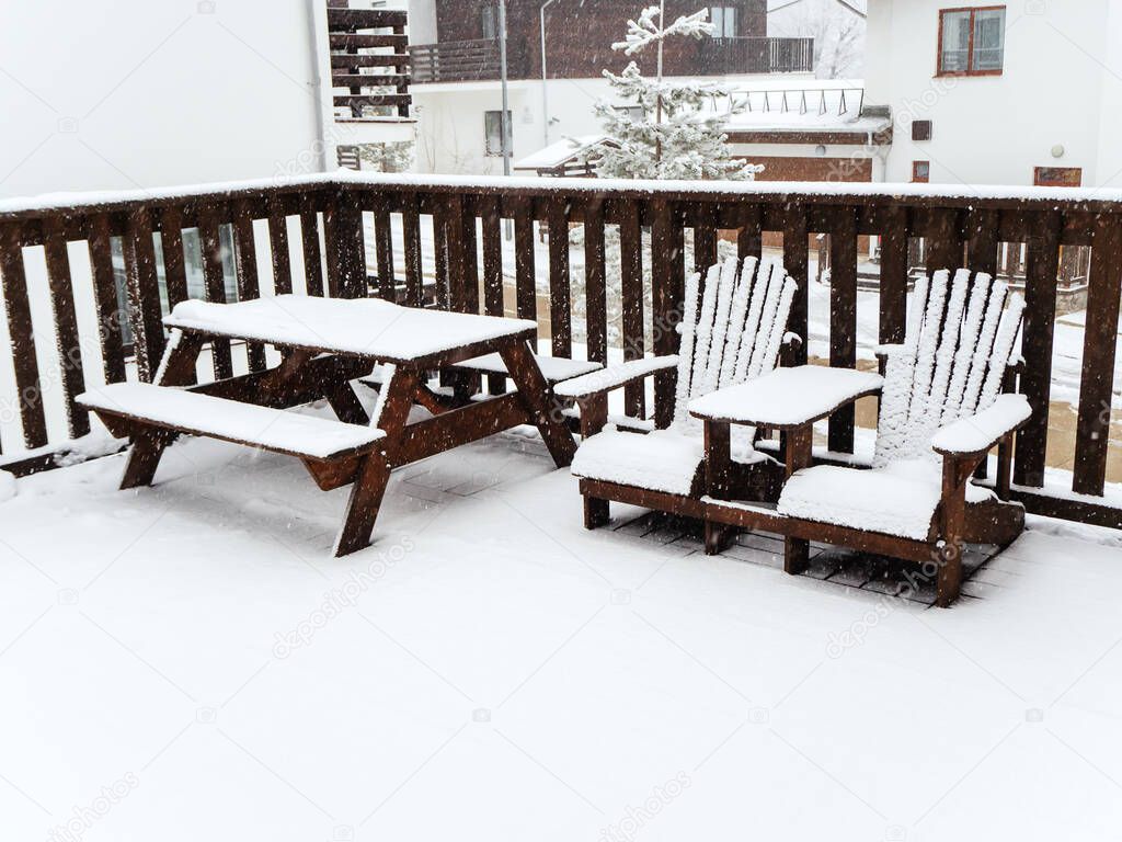 Two wooden chairs and a table with benches stand on a snow-covered veranda under a snowfall