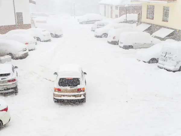 Bilen kör genom en snötäckt gård i en snöstorm förbi parkerade bilar täckta med snö — Stockfoto