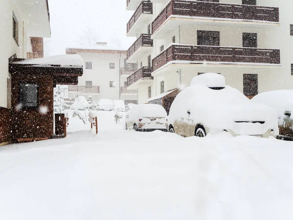 Snöig väg med bilar täckta med snö på stugornas bakgrund. Snöfall i skidorten — Stockfoto