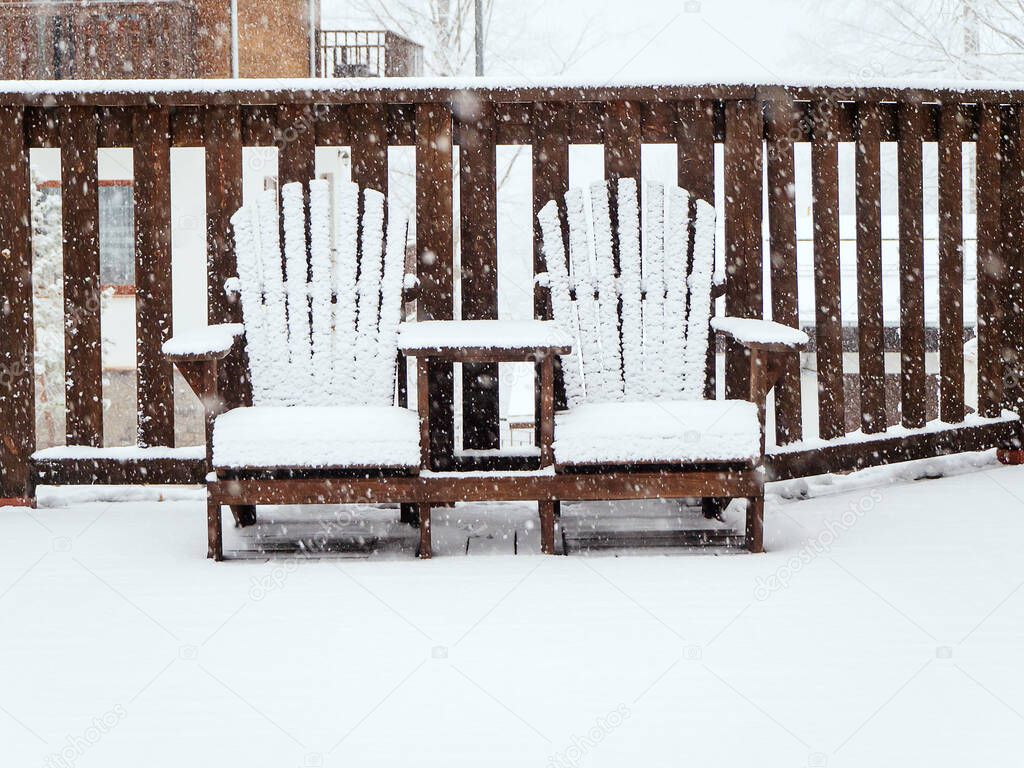 Two wooden garden chairs sit on a terrace covered with snow in winter during snowfall