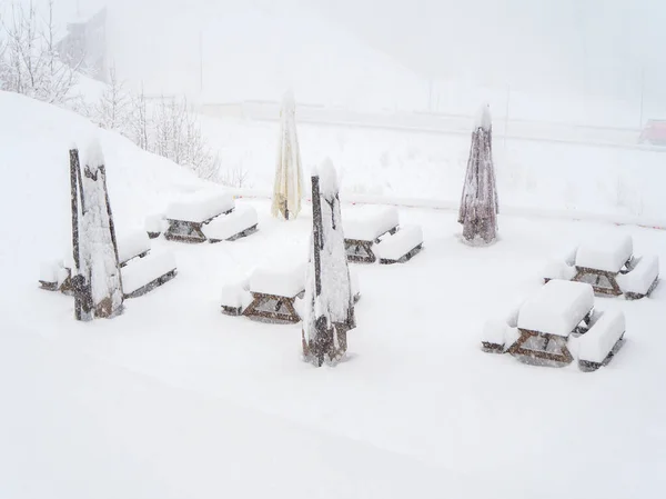 Snow-covered tables with benches and folded outdoor umbrellas stand on the site on a winter day during a blizzard — Stock Photo, Image