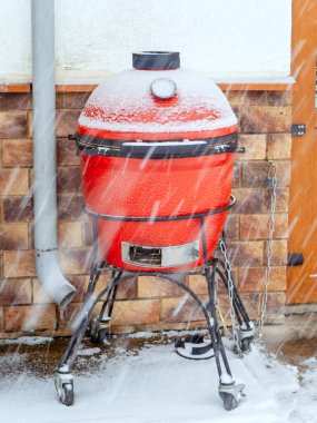 Ceramic red barbecue grill stands outside chained to the wall in snowy weather clipart