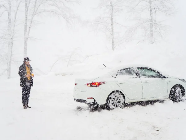 En man i hatt står bakom en vit bil under en tung snöfall på en vinterdag — Stockfoto