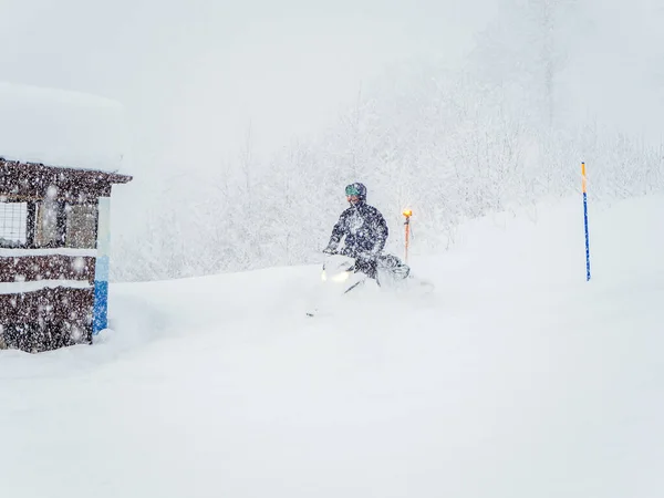 Um homem em um snowmobile no tempo nebuloso nevado dirige até a cabana — Fotografia de Stock