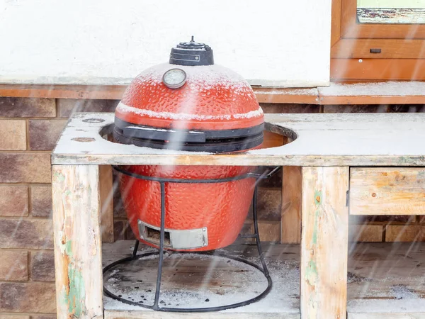 Red ceramic barbecue grill stands outside in the hole of a wooden tabletop on a snowy winter day — Stock Photo, Image