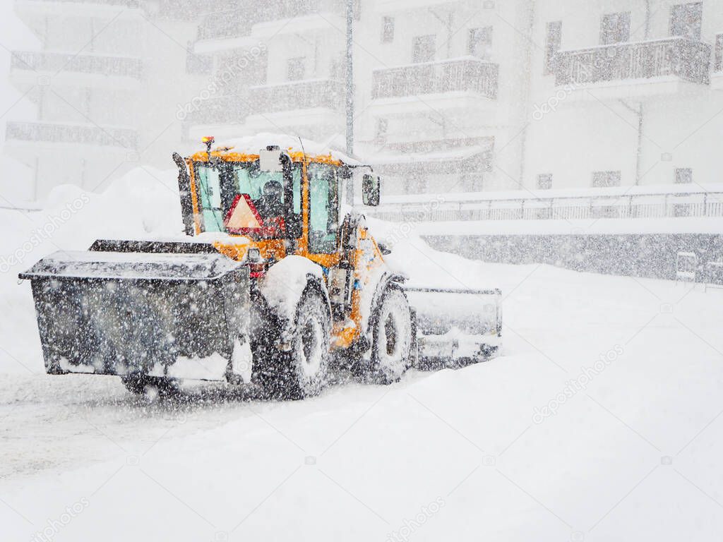 Tractor clears snow from the road in front of the building during heavy snowfall and overcast