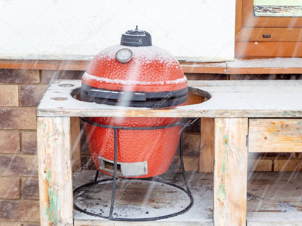 Red ceramic barbecue grill stands outside in the hole of a wooden tabletop on a snowy winter day
