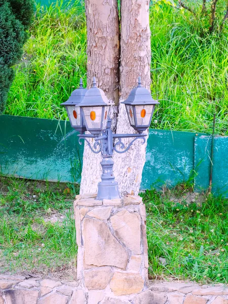 Lámpara de calle decorativa triple en un poste corto y un soporte con mampostería contra el fondo de un tronco de árbol y hierba verde — Foto de Stock