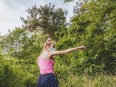 Maskeli bir kız, ormanın içinde elini önlerine uzatmış ve yumruğunu sıkarak duruyor. Salgın süresince spor.