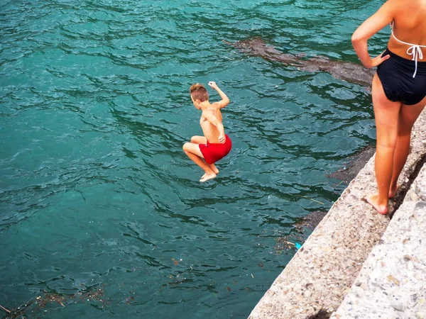 Niño en traje de baño rojo saltando al mar con las piernas dobladas —  Fotos de Stock