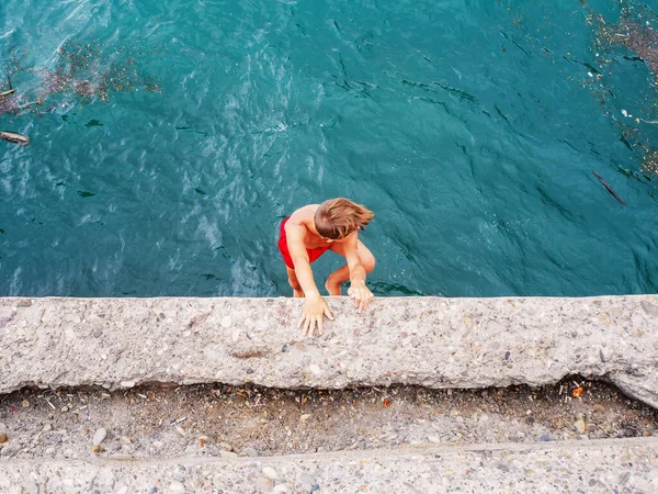 Un ragazzo in costume da bagno rosso scende da un molo di cemento nell'acqua del mare — Foto Stock
