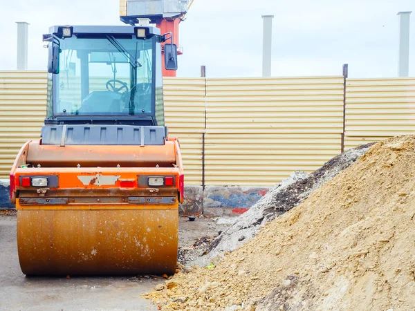 Válec na překupování asfaltu stojí u hromady písku na pozadí stavebního plotu — Stock fotografie