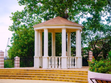 Beyaz kolonları olan bir çardak, şehir parkında yeşil ağaçların altında duruyor.