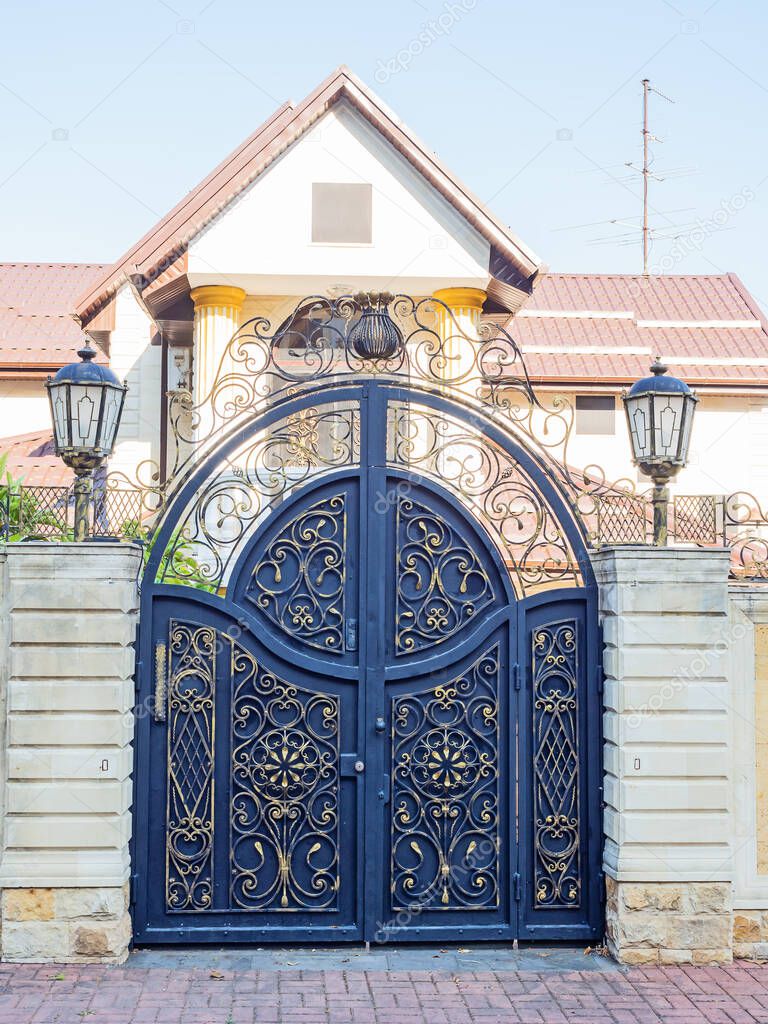 Rounded iron gates with a gilded wrought pattern and a fence enclosing a cottage